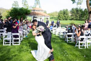 Groom dips bride for a kiss during outdoor wedding ceremony with guests seated on white chairs.