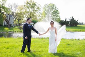 Bride and groom holding hands on green grass with flowing wedding veil and pond backdrop.
