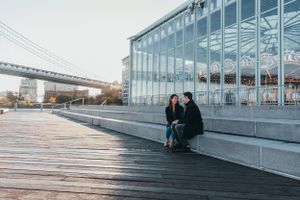 A couple sitting together on a waterfront boardwalk near a glass building with a bridge visible in the background.