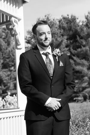 Black and white portrait of groom in dark suit smiling beside white gazebo outdoors.