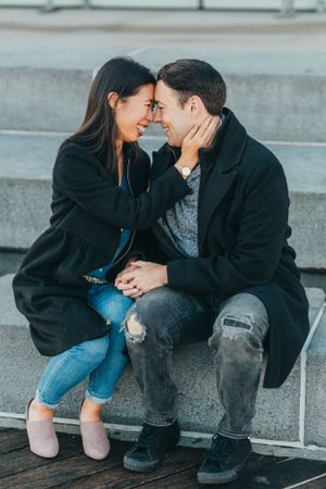 A couple sitting on steps, smiling and touching foreheads romantically, both wearing black coats in an outdoor urban setting.