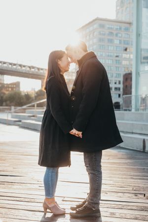 A romantic couple holding hands and touching foreheads on a sunny urban boardwalk with city buildings behind them.