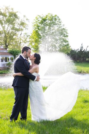 Bride and groom sharing a romantic kiss with the bride's veil flowing in the wind by a pond.