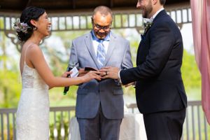 Bride and groom exchange rings with officiant during outdoor wedding ceremony under a gazebo.