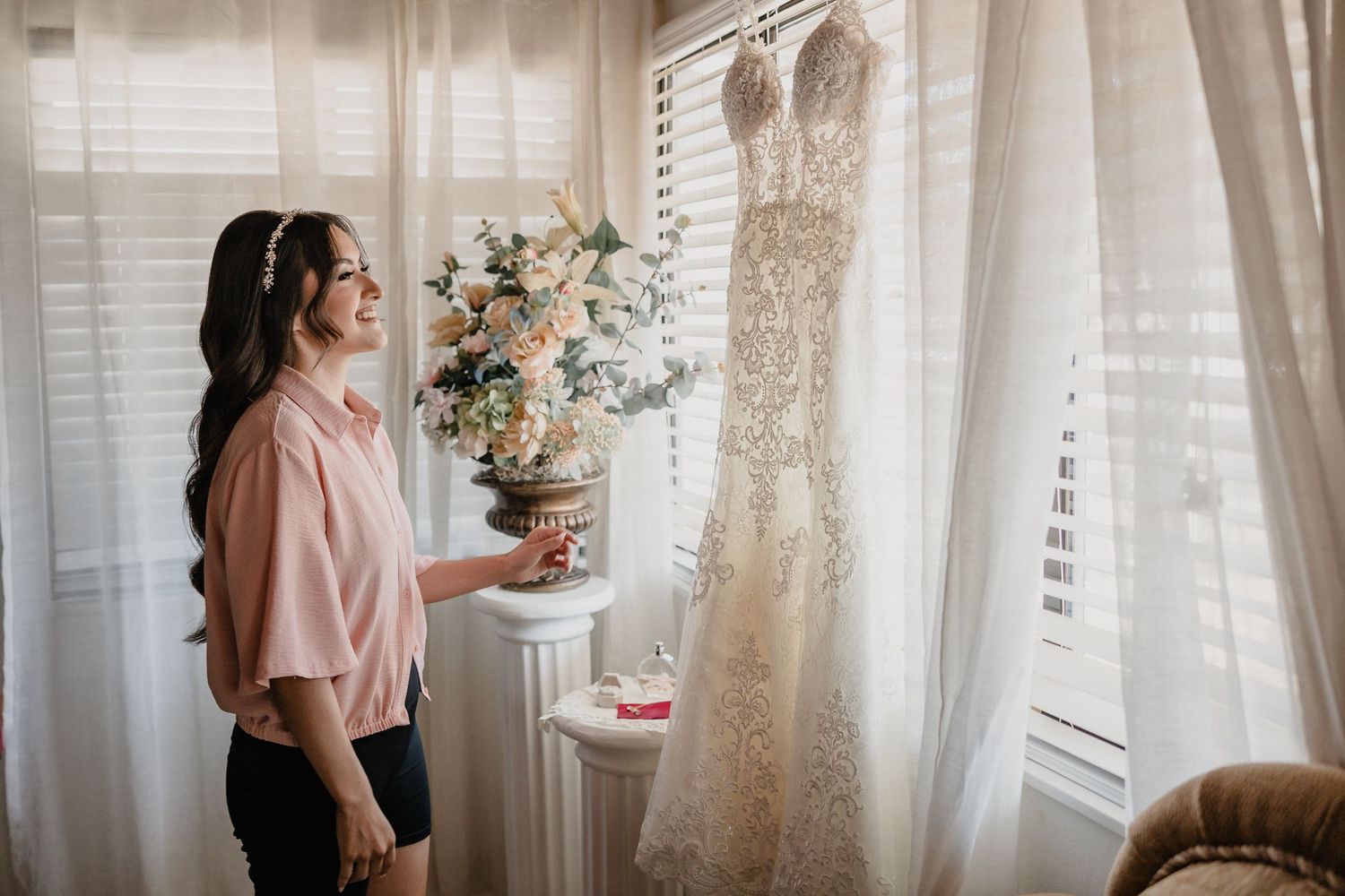 Woman in pink blouse admiring elegant lace wedding dress hanging by window.