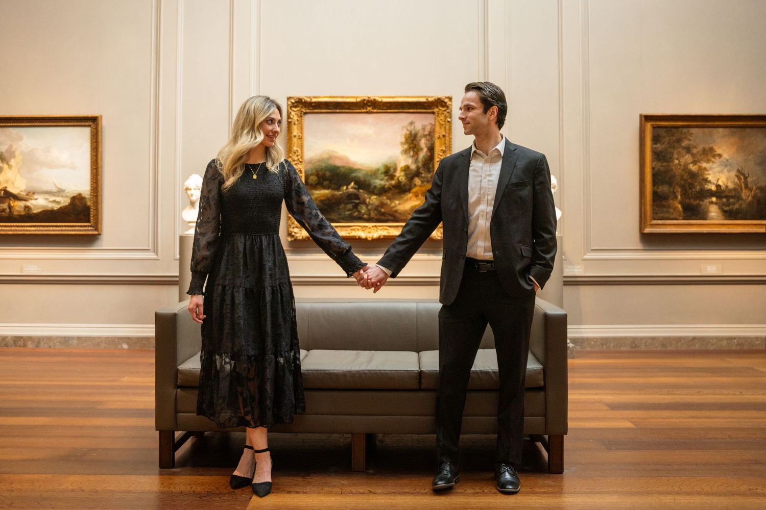 Two people sit together on a wooden bench in an art gallery displaying landscape paintings on the walls.