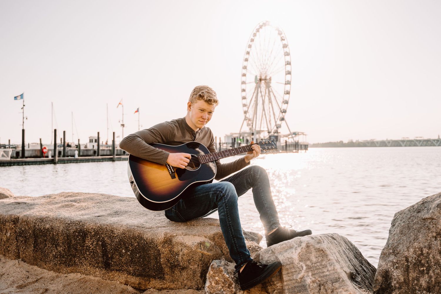 Person in casual attire sits on rocky seawall with acoustic guitar at sunset overlooking industrial harbor.