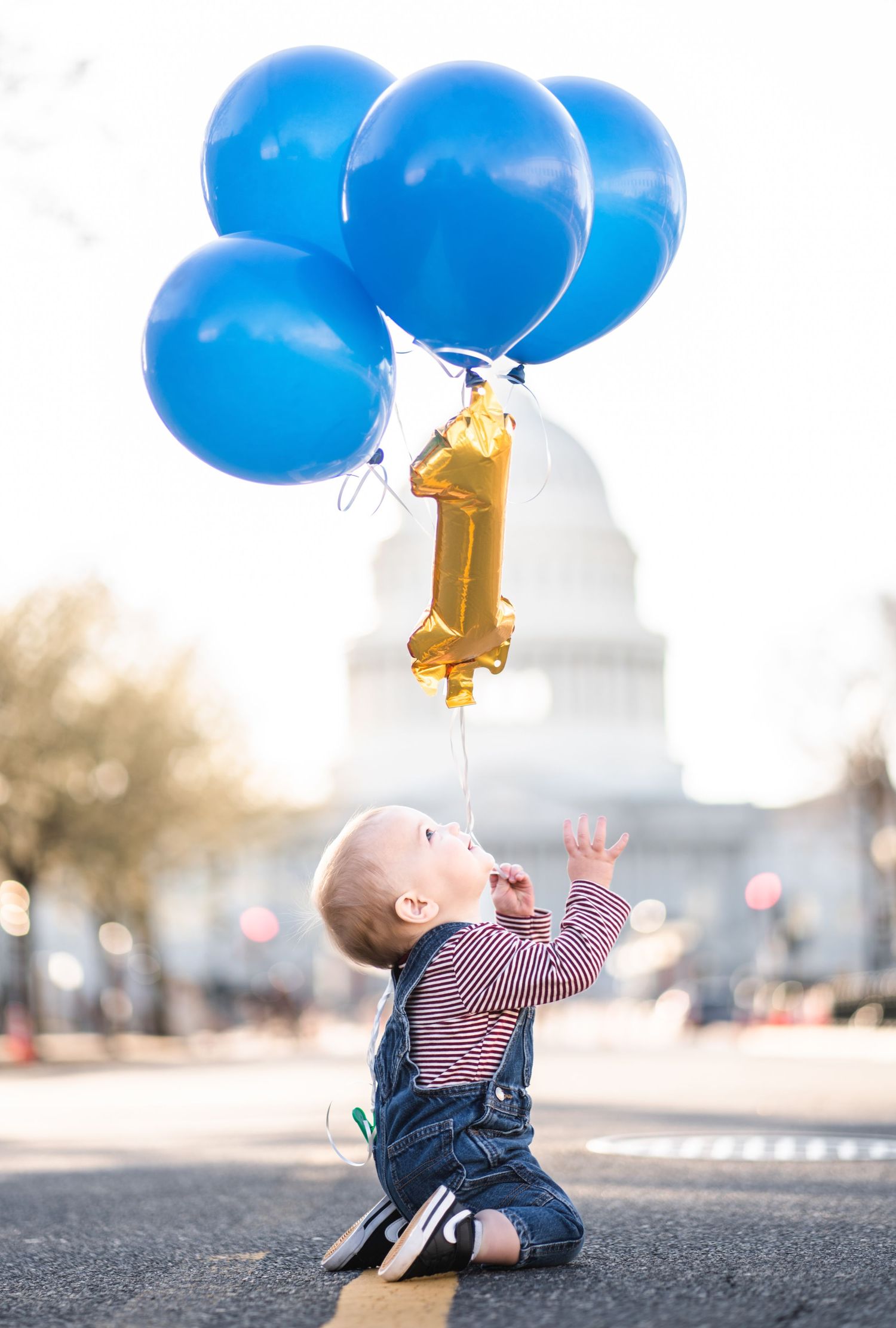 A baby in a striped shirt crawls on pavement while looking up at floating blue and gold birthday balloons.