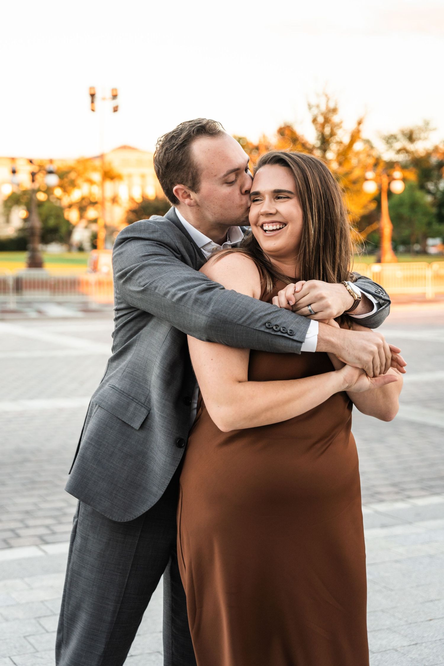 A couple shares a joyful embrace outdoors during sunset, with the person in grey suit hugging from behind.