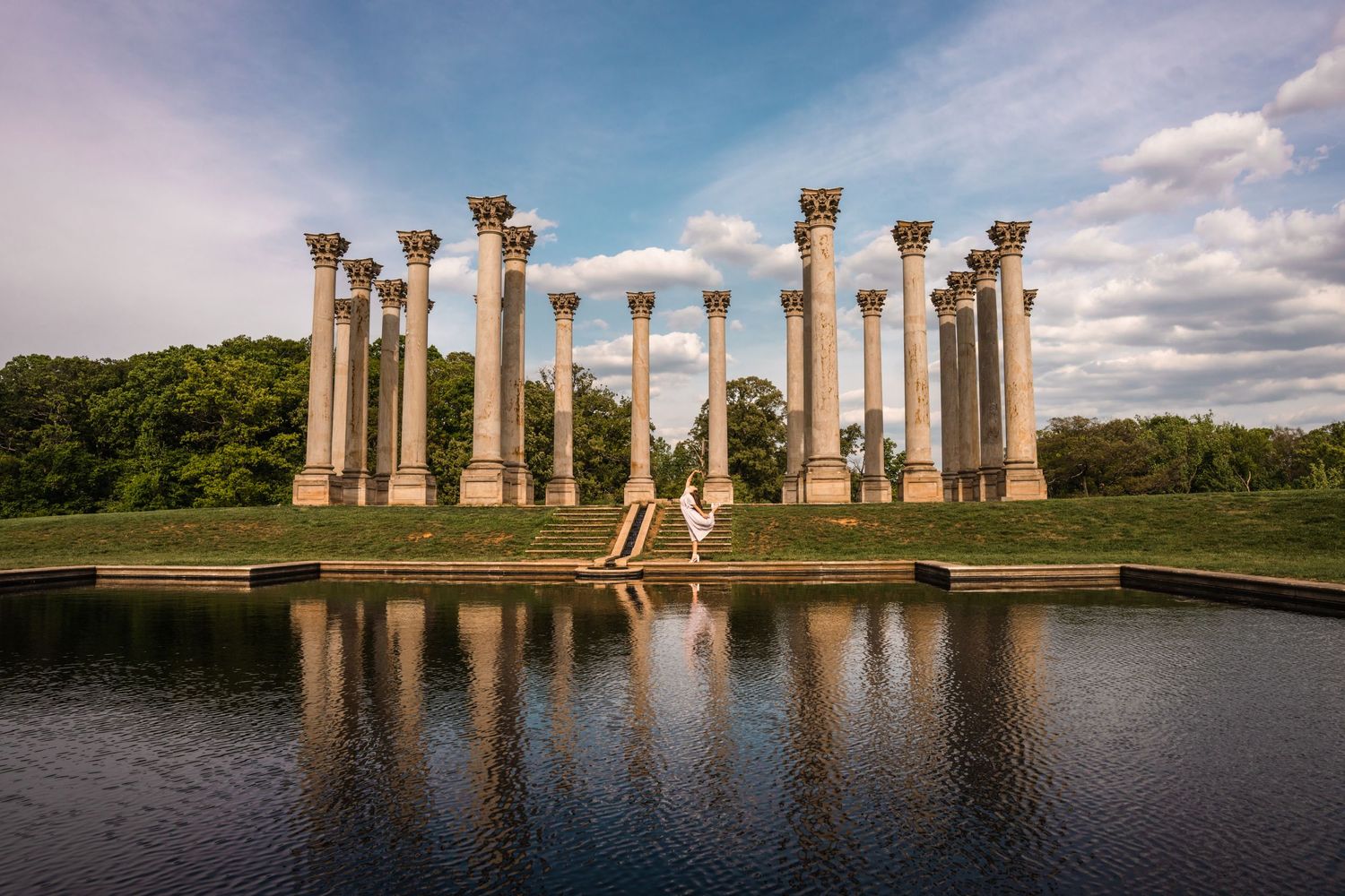 Ancient stone columns stand reflected in calm water at sunset against a dramatic cloudy sky.