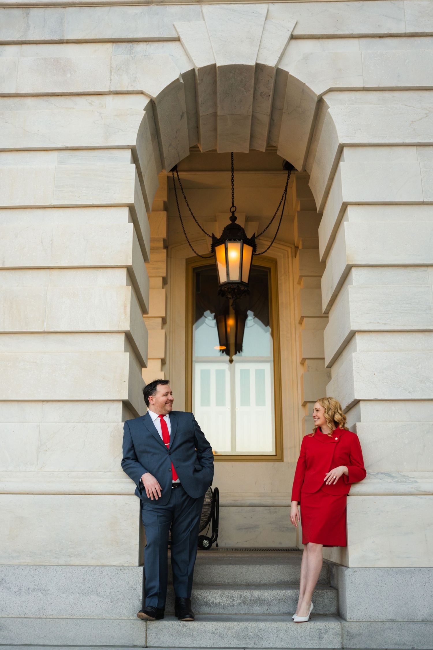 Two people in professional attire stand beneath an ornate archway with a hanging lantern at a government building.