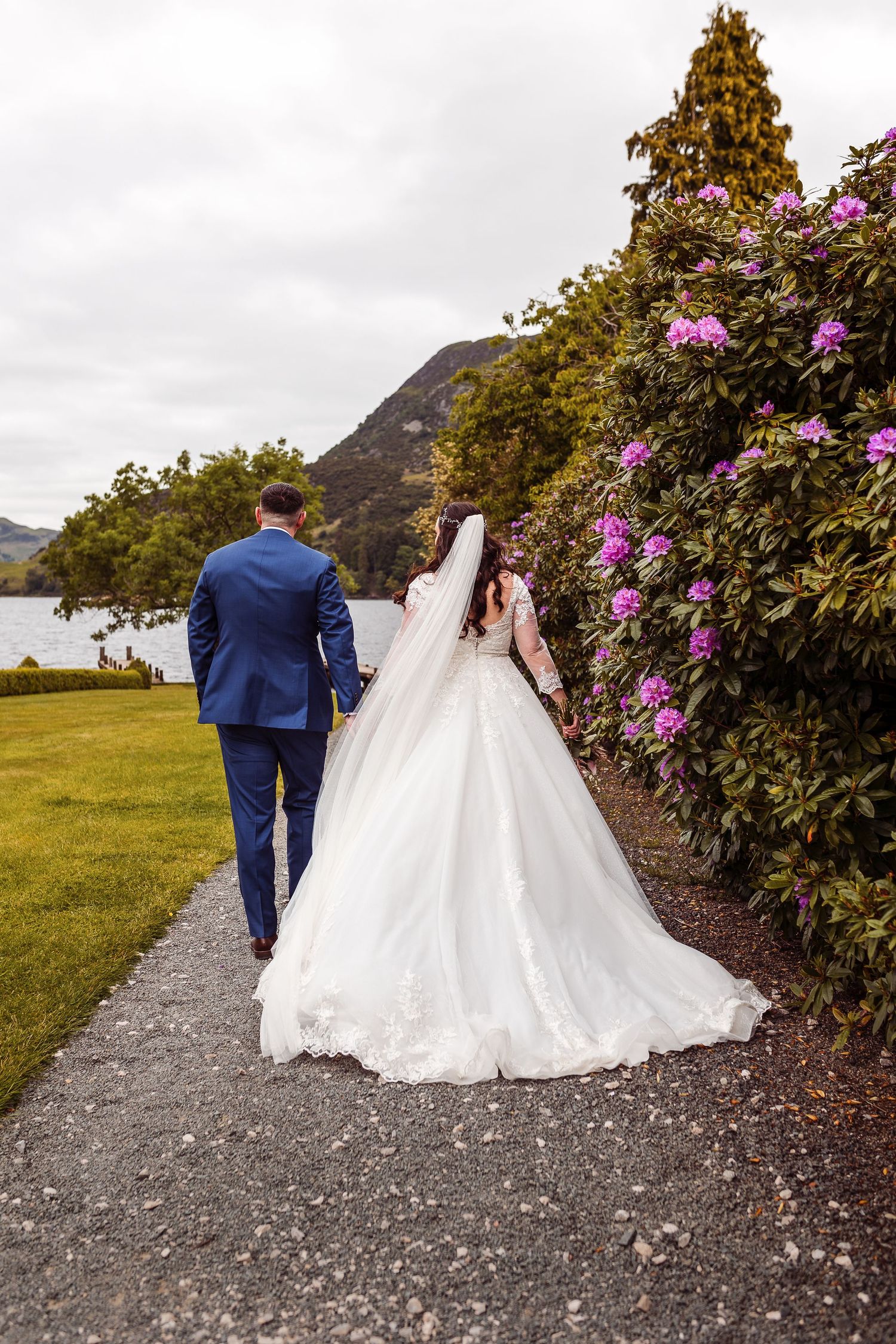 A wedding couple walks along a path bordered by blooming rhododendrons near a lake.