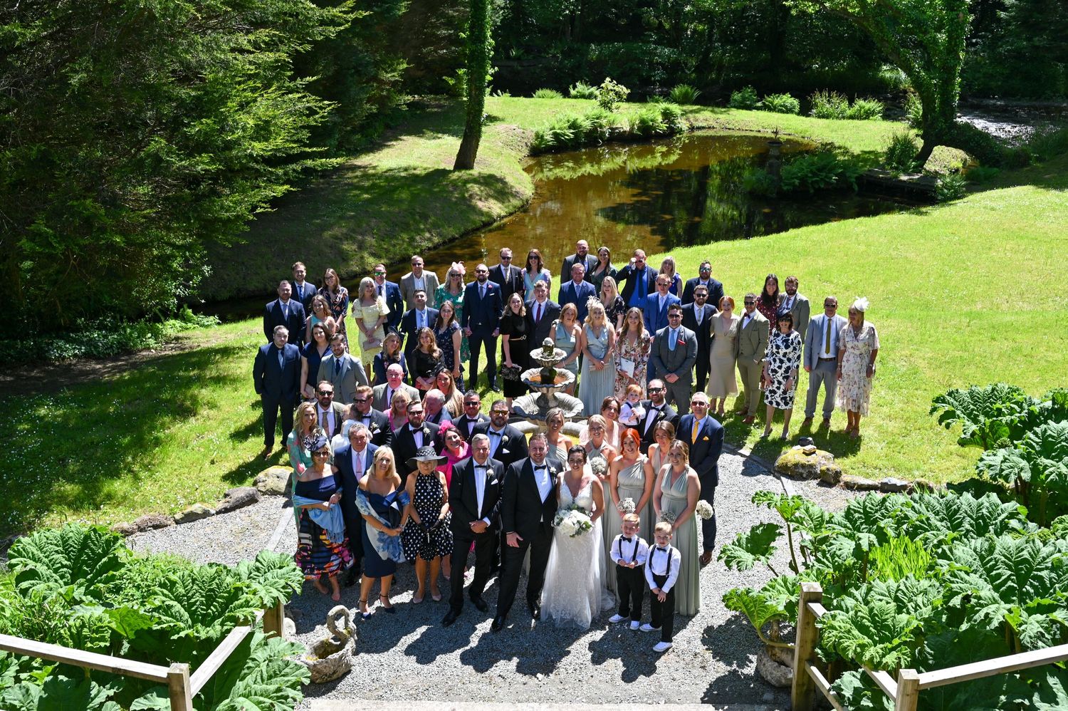 Large wedding party group photo taken on garden path surrounded by lush greenery.