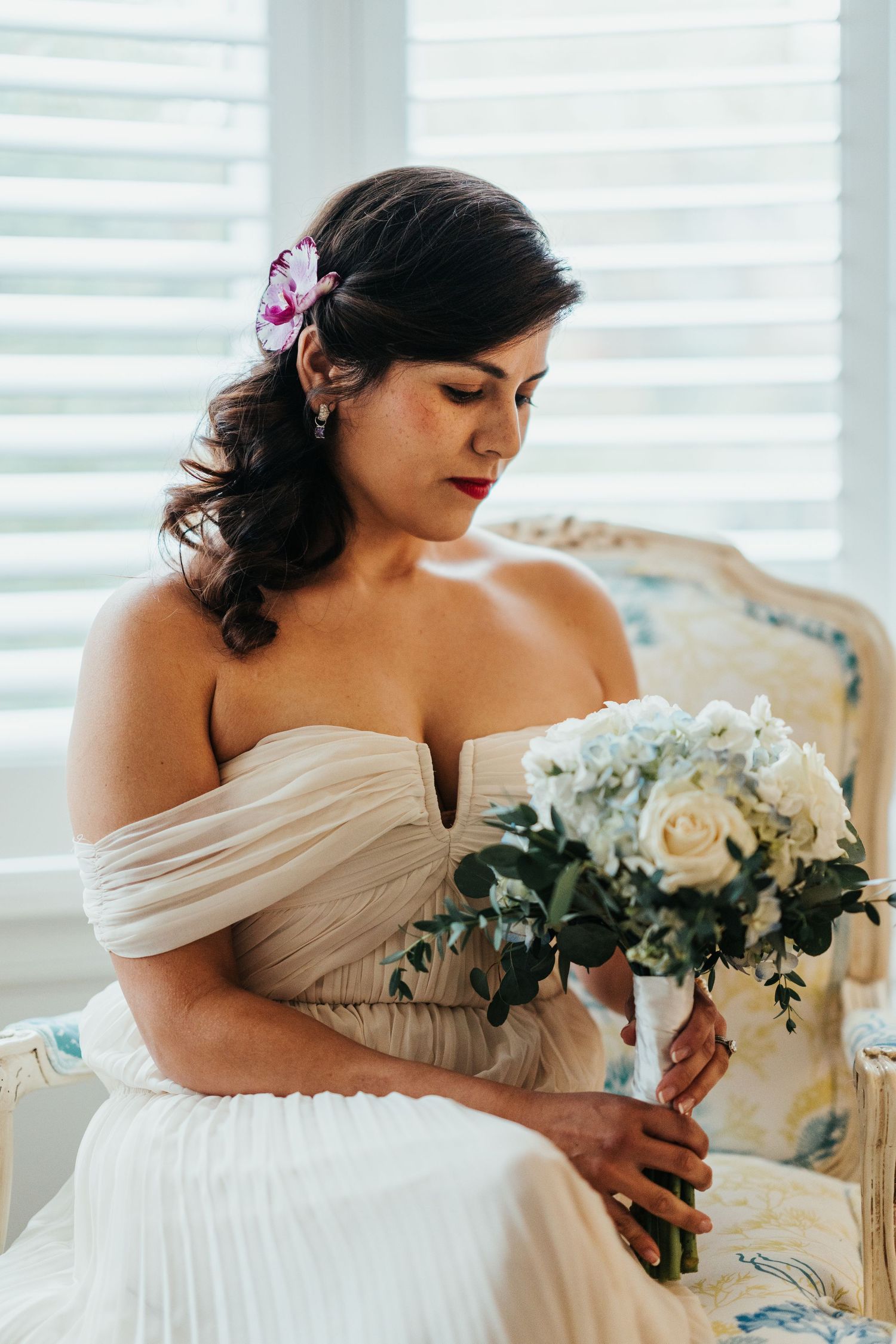 Bride in off-shoulder white dress holding elegant white rose bouquet in vintage chair.