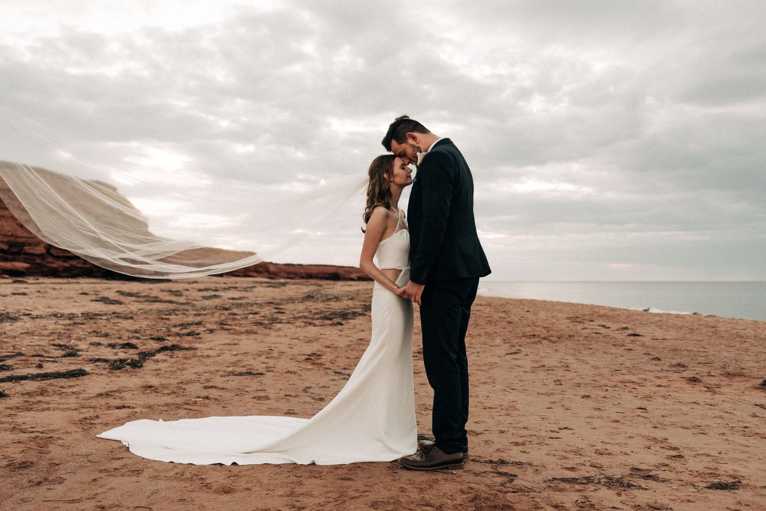 A couple shares an intimate moment on a beach with dramatic wedding dress train flowing in the wind.