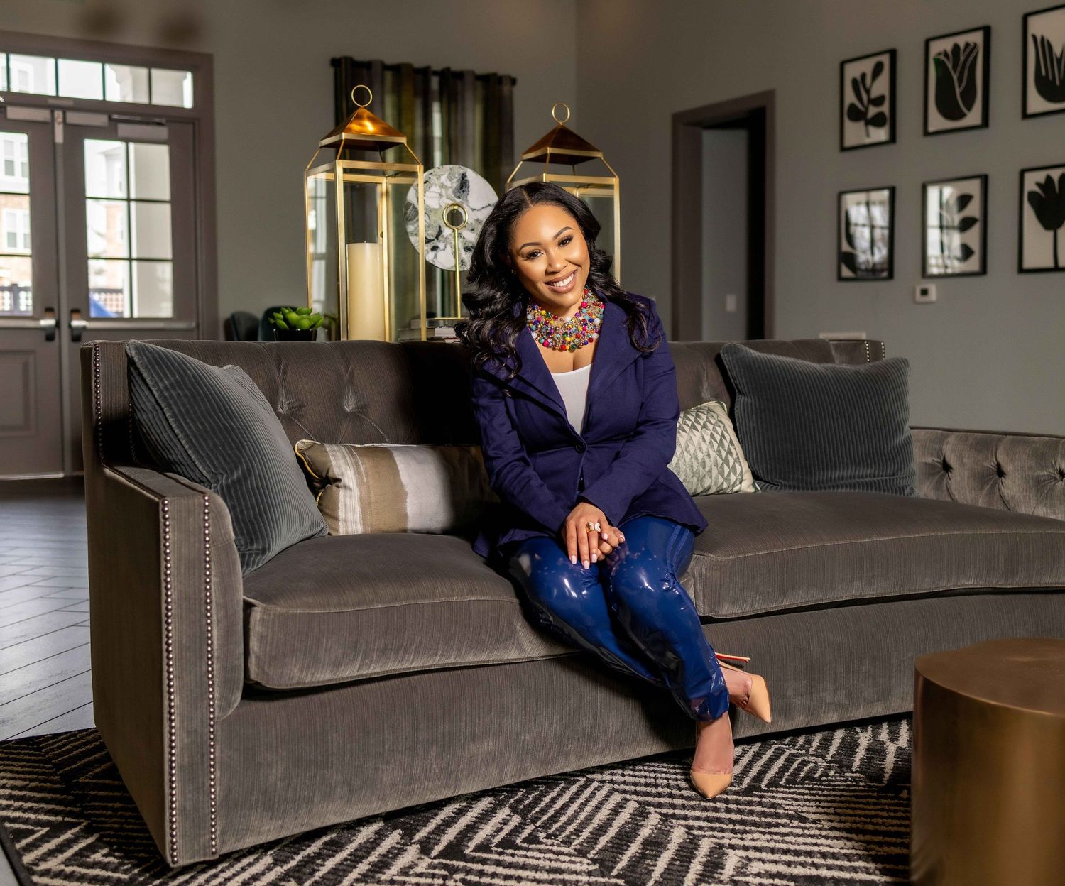 Stylish individual in navy blazer relaxing on gray velvet couch with pendant lights and wall art.