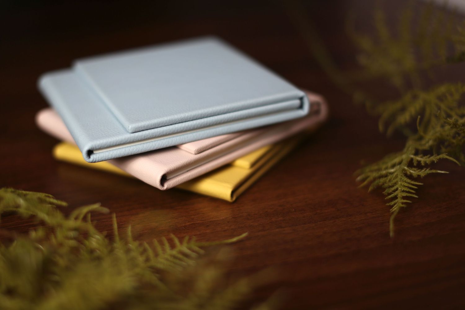 Neatly stacked photo albums with soft fern leaves on a dark wooden surface.