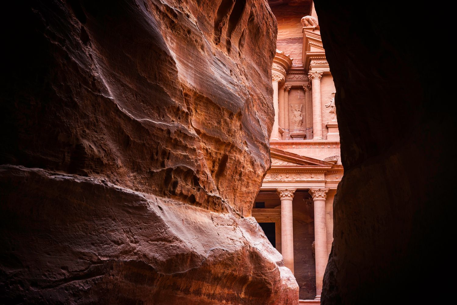Ancient carved temple facade of Petra visible through narrow red rock canyon walls in Jordan.