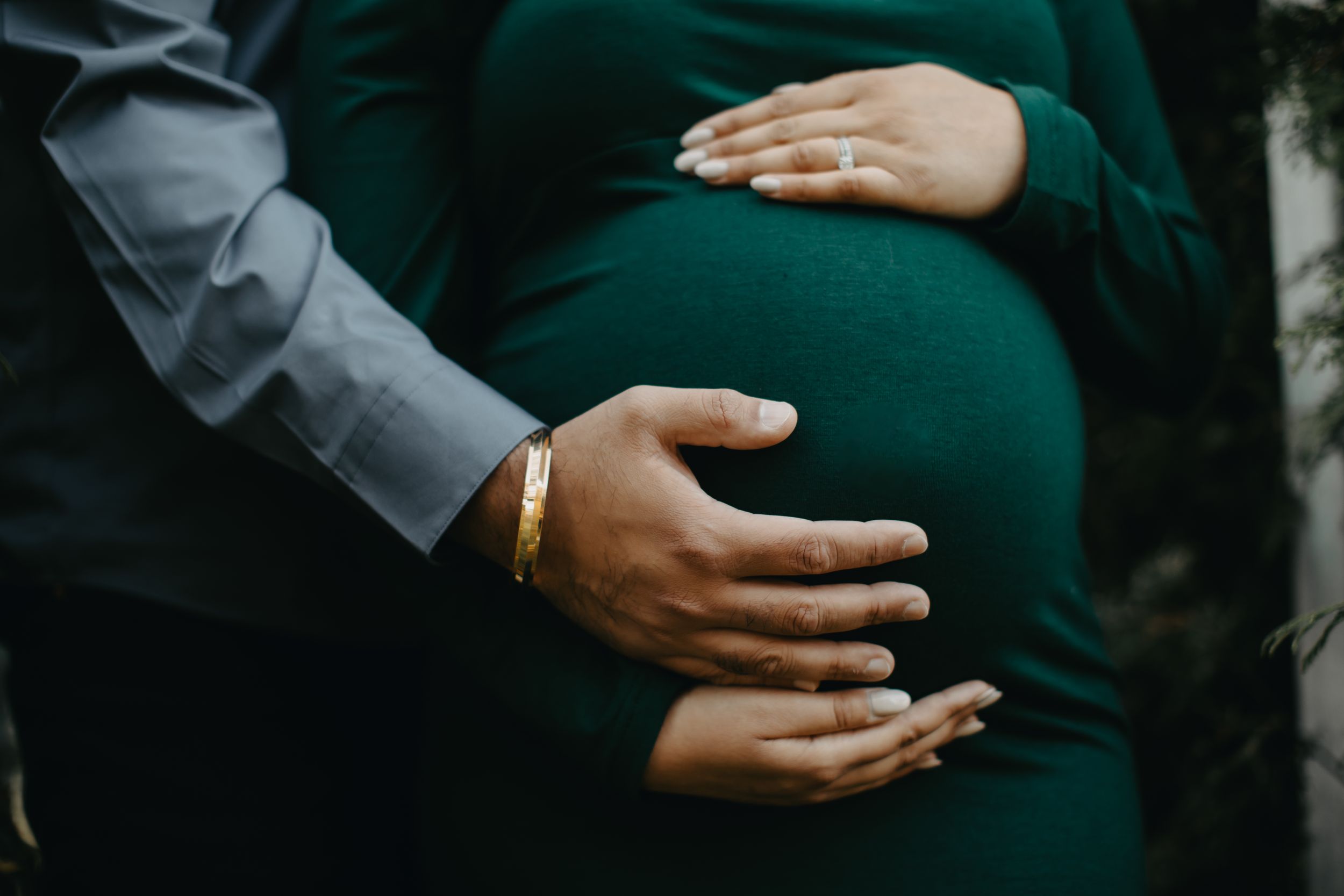 Intimate close-up of hands tenderly holding pregnant belly in dark green dress.
