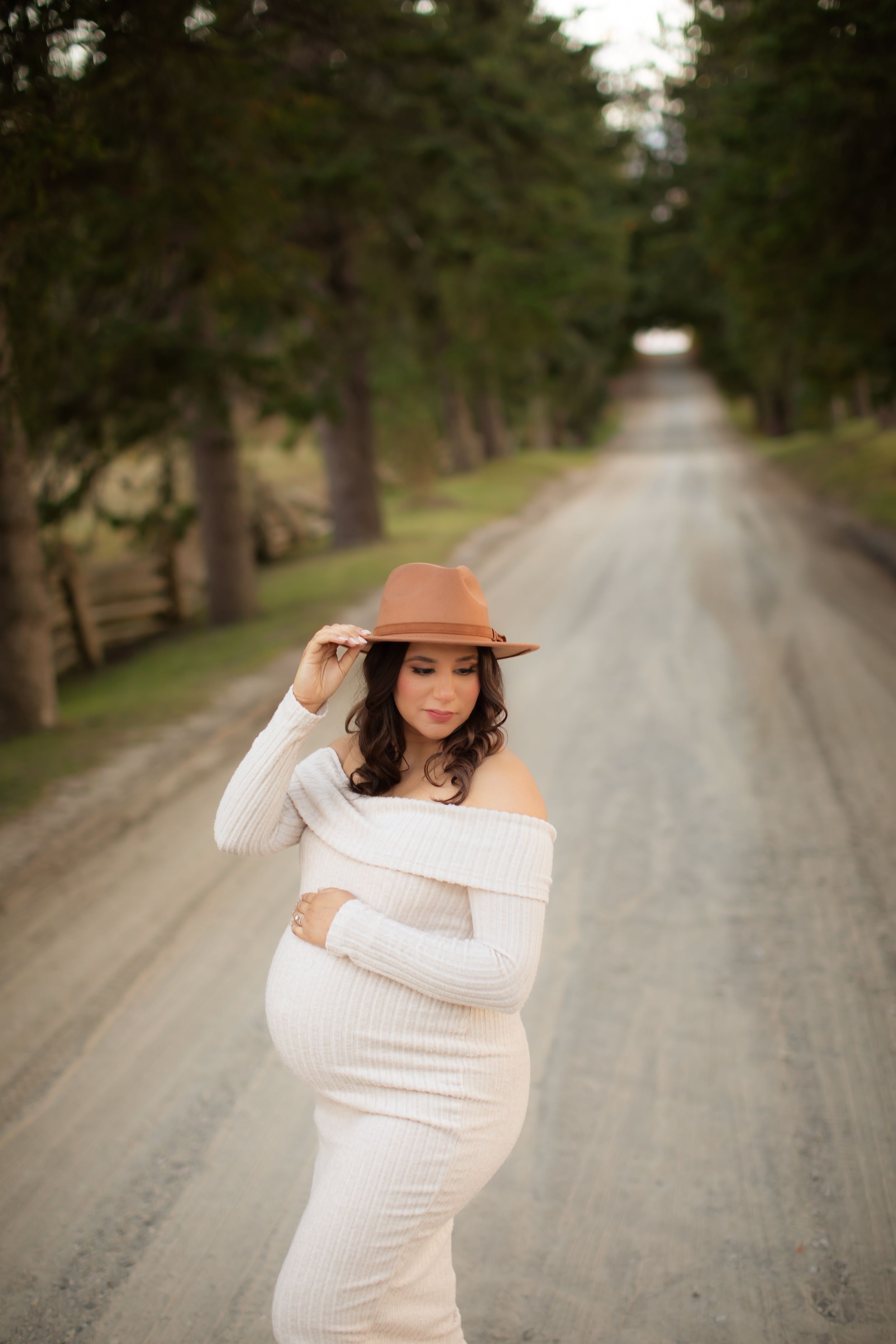 Maternity photoshoot on a tree-lined dirt road with a person in a white dress and brown hat.