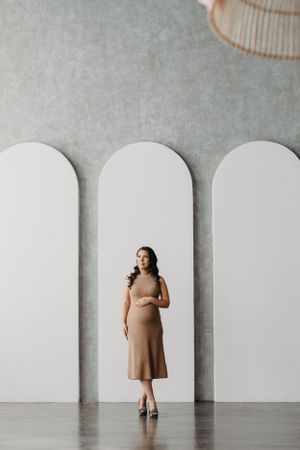 Woman in brown dress poses against white archways in minimalist studio setting.