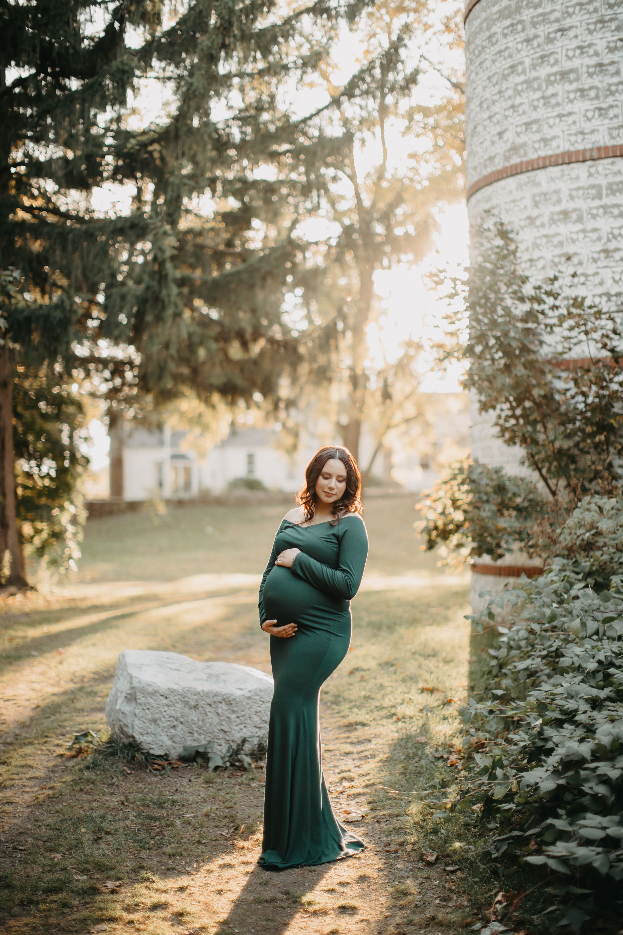 Maternity photo in emerald green dress against stone wall and autumn foliage.