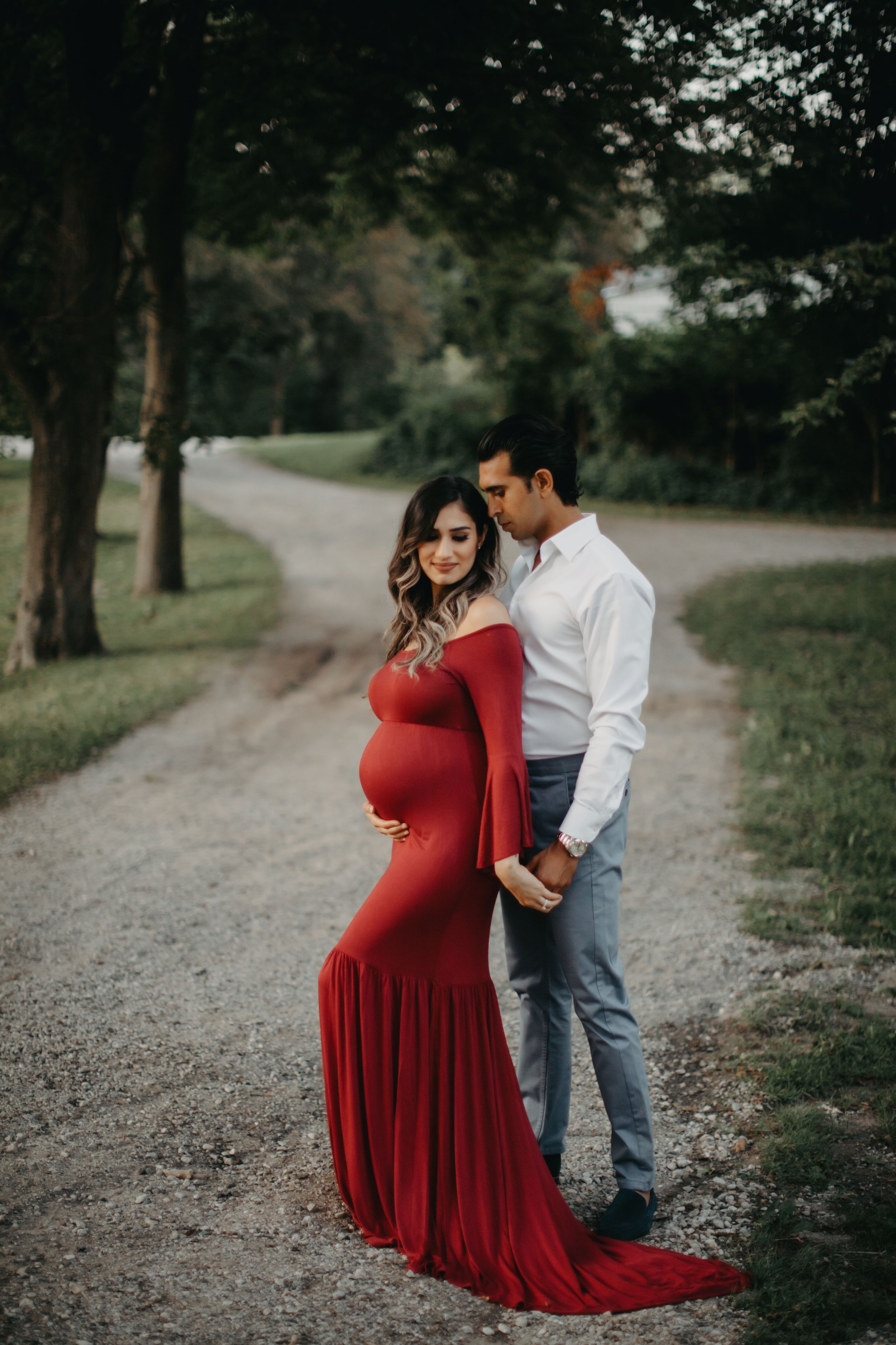 Maternity photoshoot on wooded path featuring couple with woman in flowing red dress.
