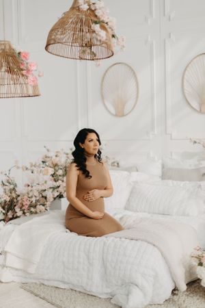 Dreamy maternity photos on white bedding with soft pink floral arrangements and birdcage decor.