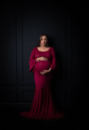 Dramatic maternity portraits against black wall featuring woman in burgundy dress with bell sleeves.