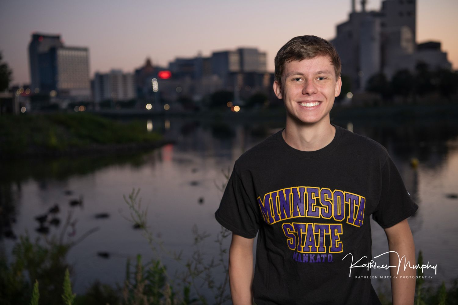 Person wearing Minnesota State t-shirt standing by river at dusk with city skyline behind.