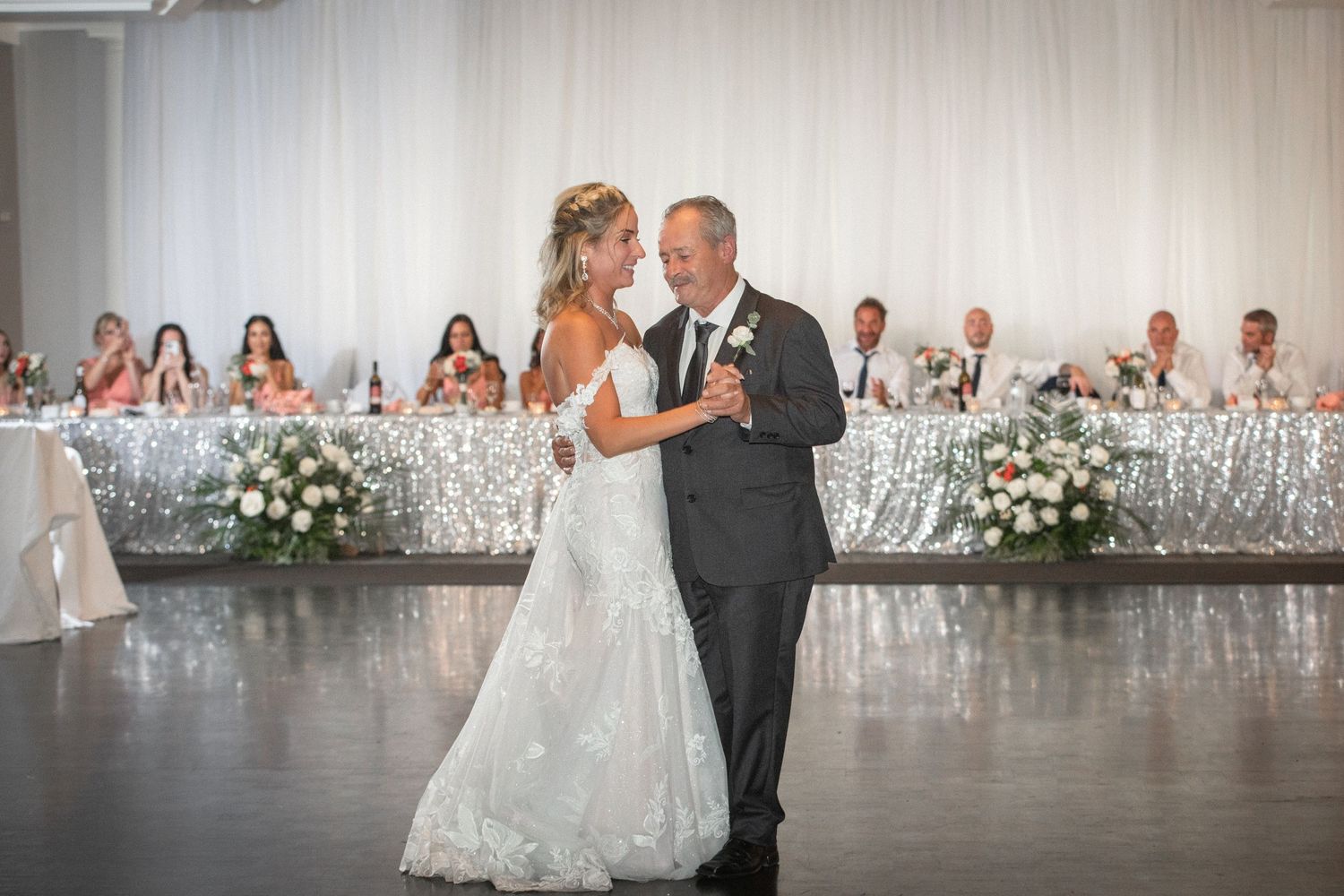 A couple shares their first dance in an elegant white reception hall decorated with floral arrangements.