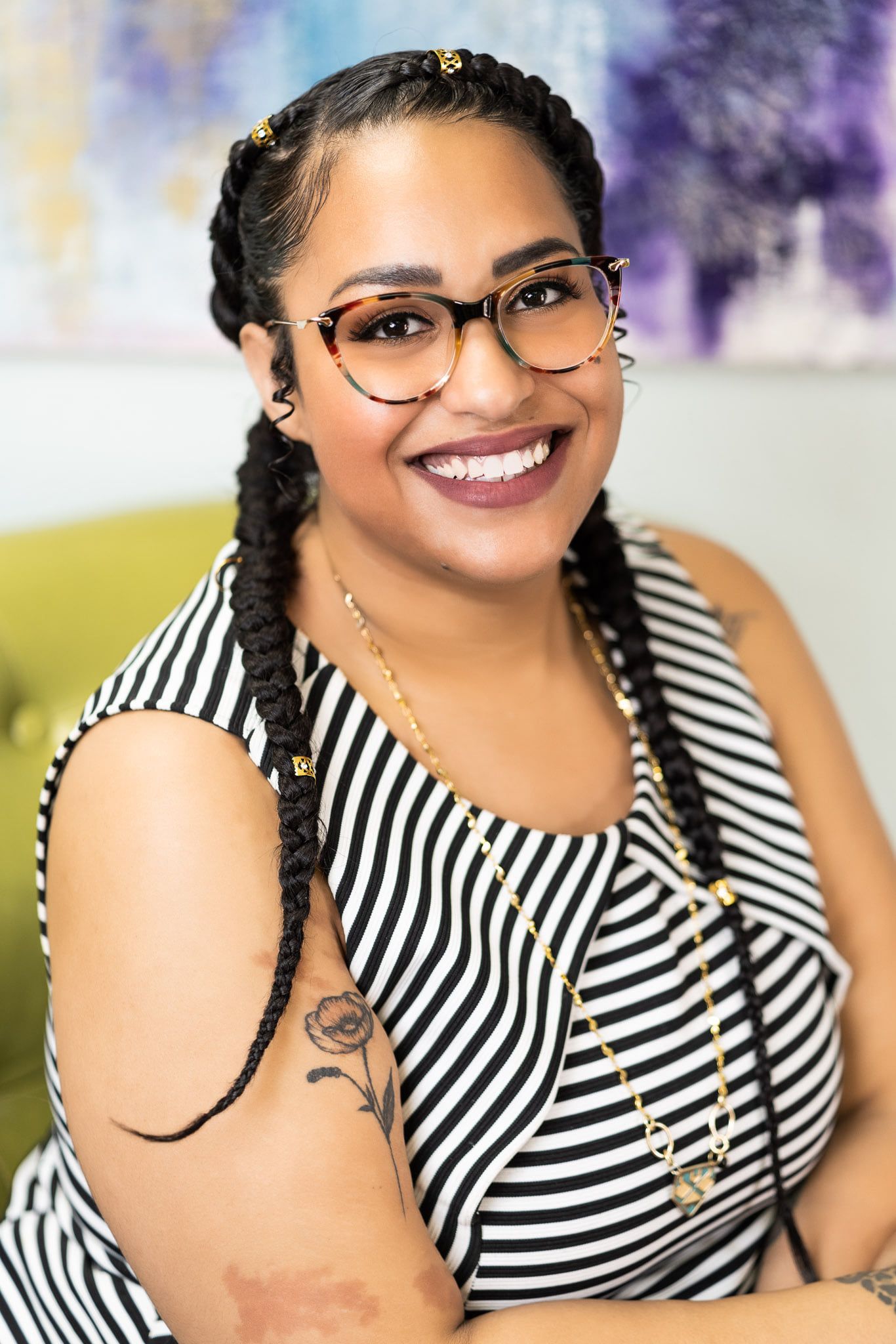 Professional portrait showing a warm smile, braided hair, glasses and striped top against colorful background.