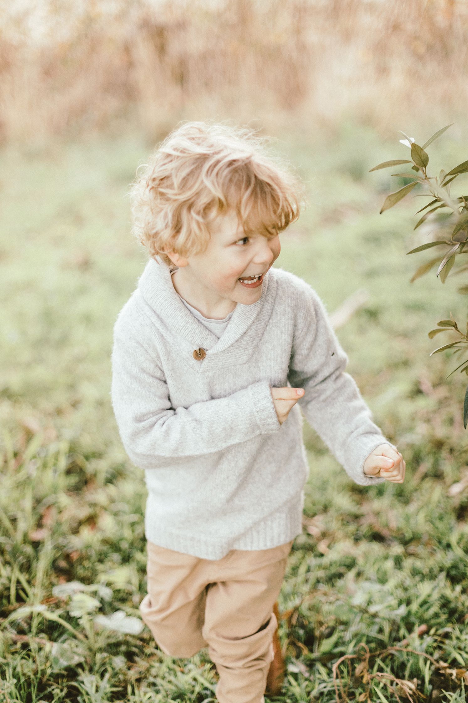 Toddler in gray sweater laughing while playing outdoors in green foliage.