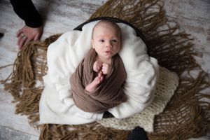 A newborn baby wrapped in brown fabric lies on a white cushion with textured backdrop.