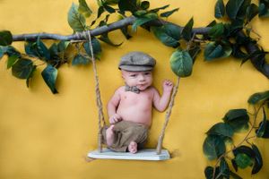 A baby wearing a cap sits on a decorative swing against a bright yellow wall with greenery.