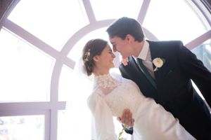 A romantic wedding couple share an intimate moment under an arched window with streaming sunlight.