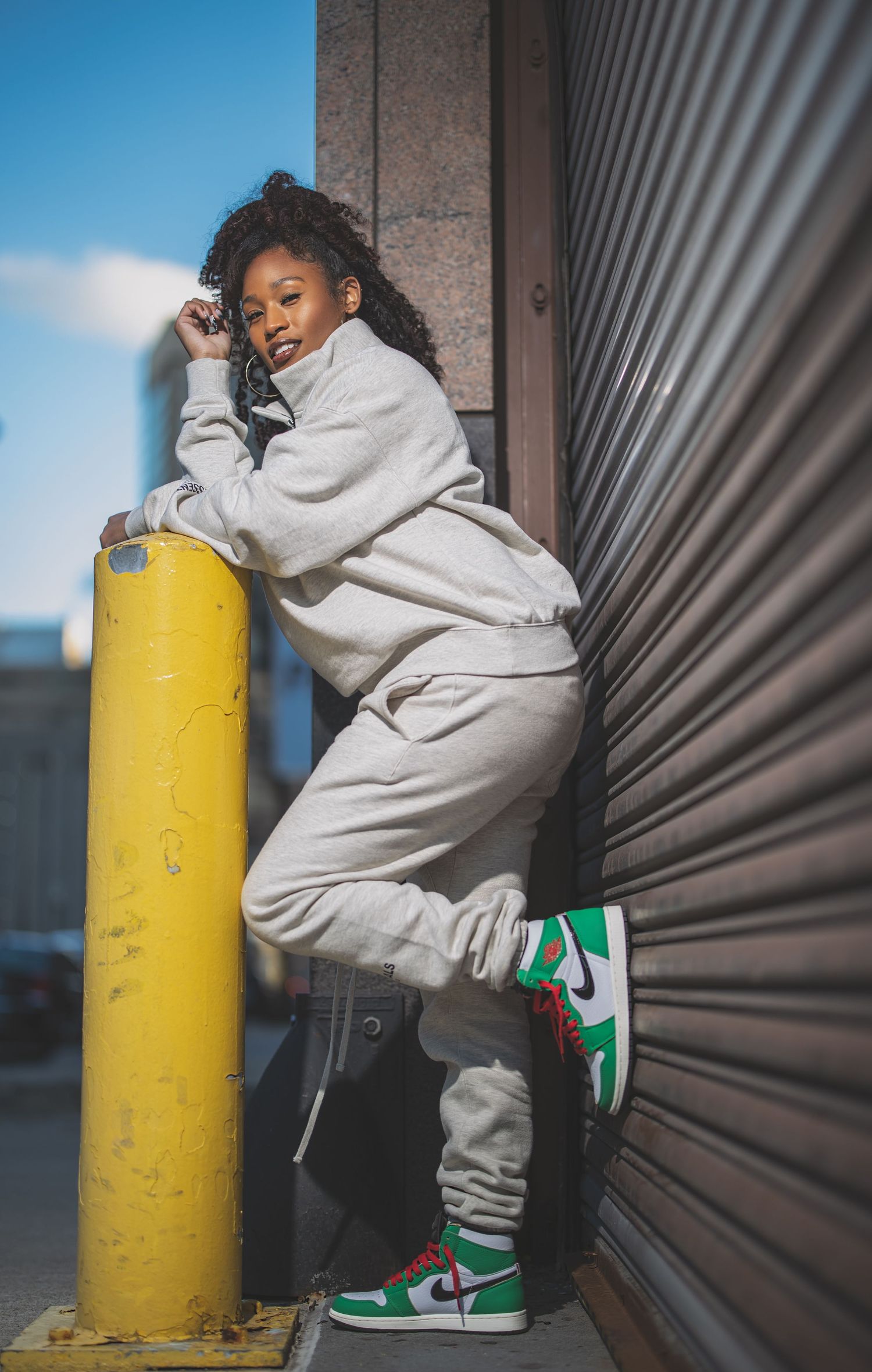 Casual streetwear pose against metal wall with yellow security pole in urban setting.