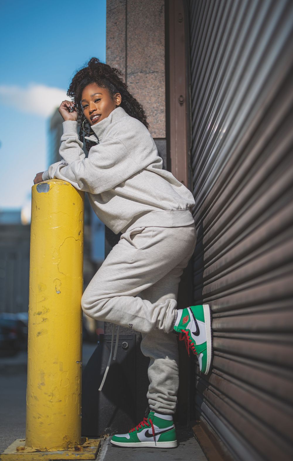 Casual streetwear pose against metal wall with yellow security pole in urban setting.