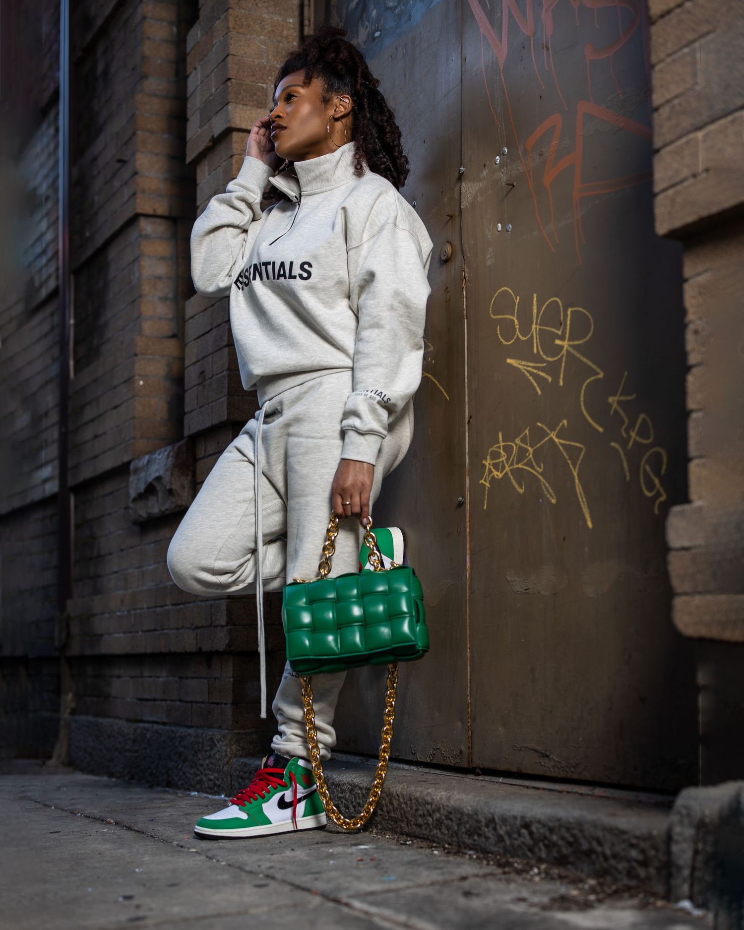 Urban fashion portrait showing cream tracksuit and green handbag against brick wall with graffiti.