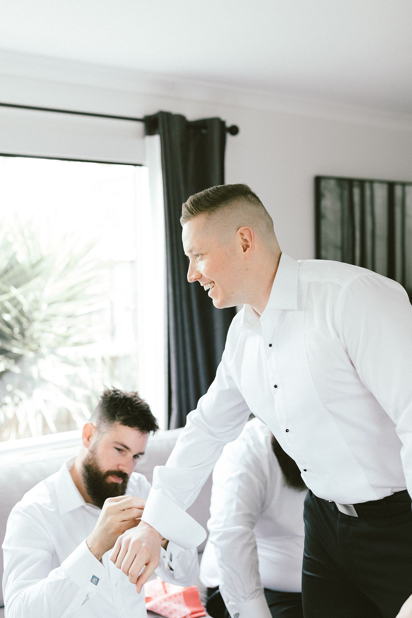 Two people in white dress shirts preparing in a room with dark curtains and natural light.