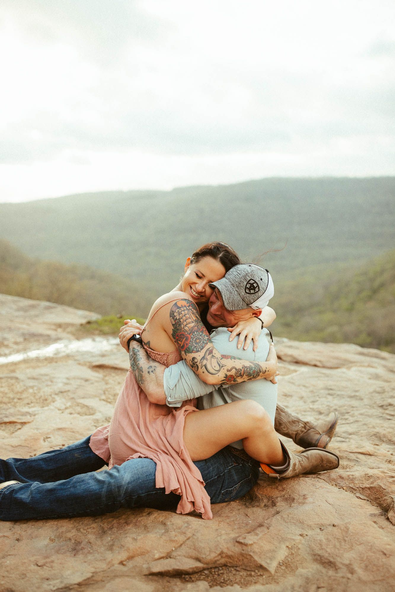 A couple shares an intimate embrace while sitting on a rocky cliff overlooking mountain valleys.