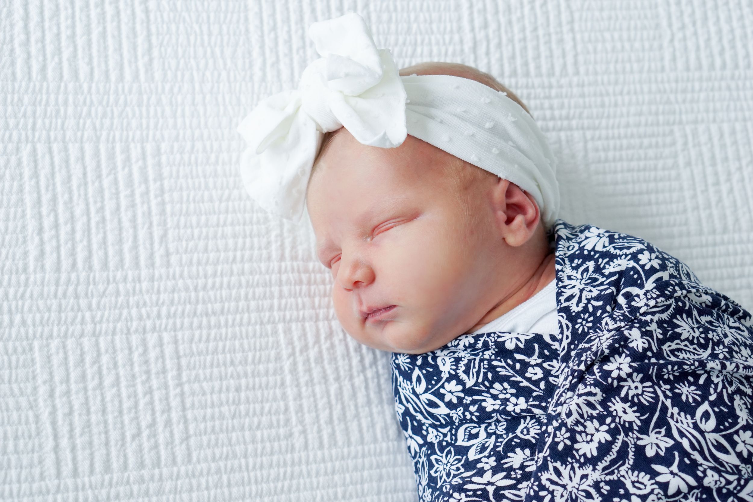 A sleeping newborn wearing a navy floral swaddle and white bow headband on white textured bedding.
