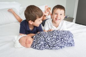 Two young children lying next to a swaddled infant on white bedding.