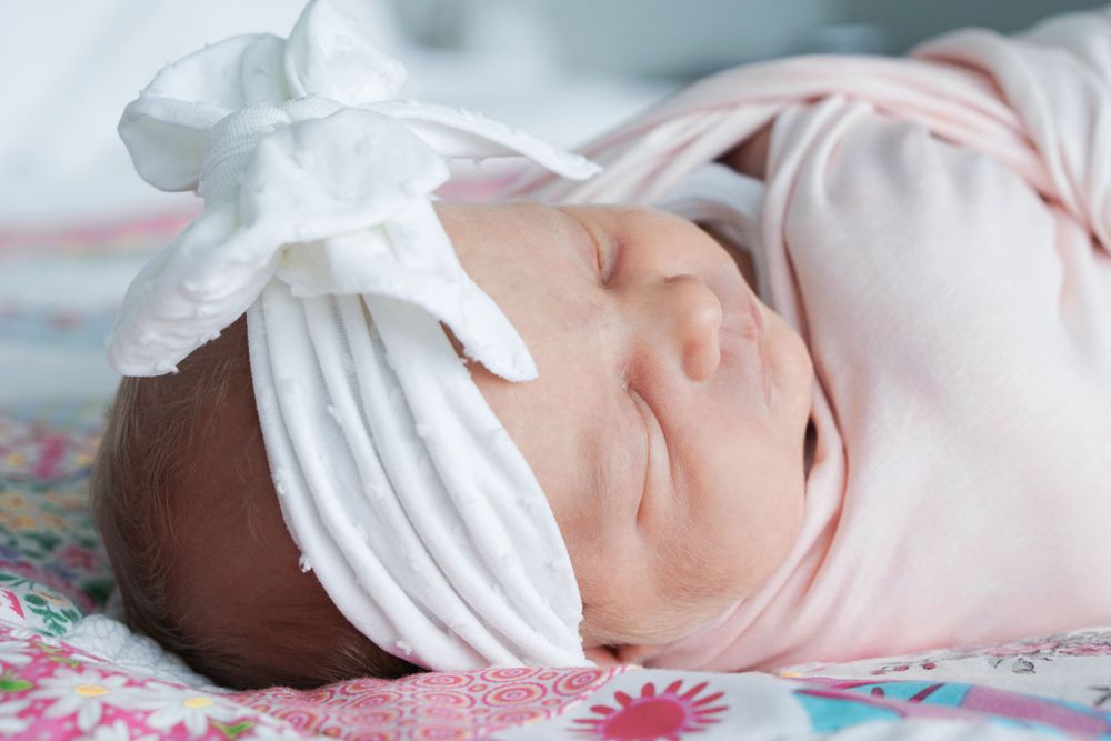 A peaceful sleeping newborn wearing a white bow headband against floral bedding.