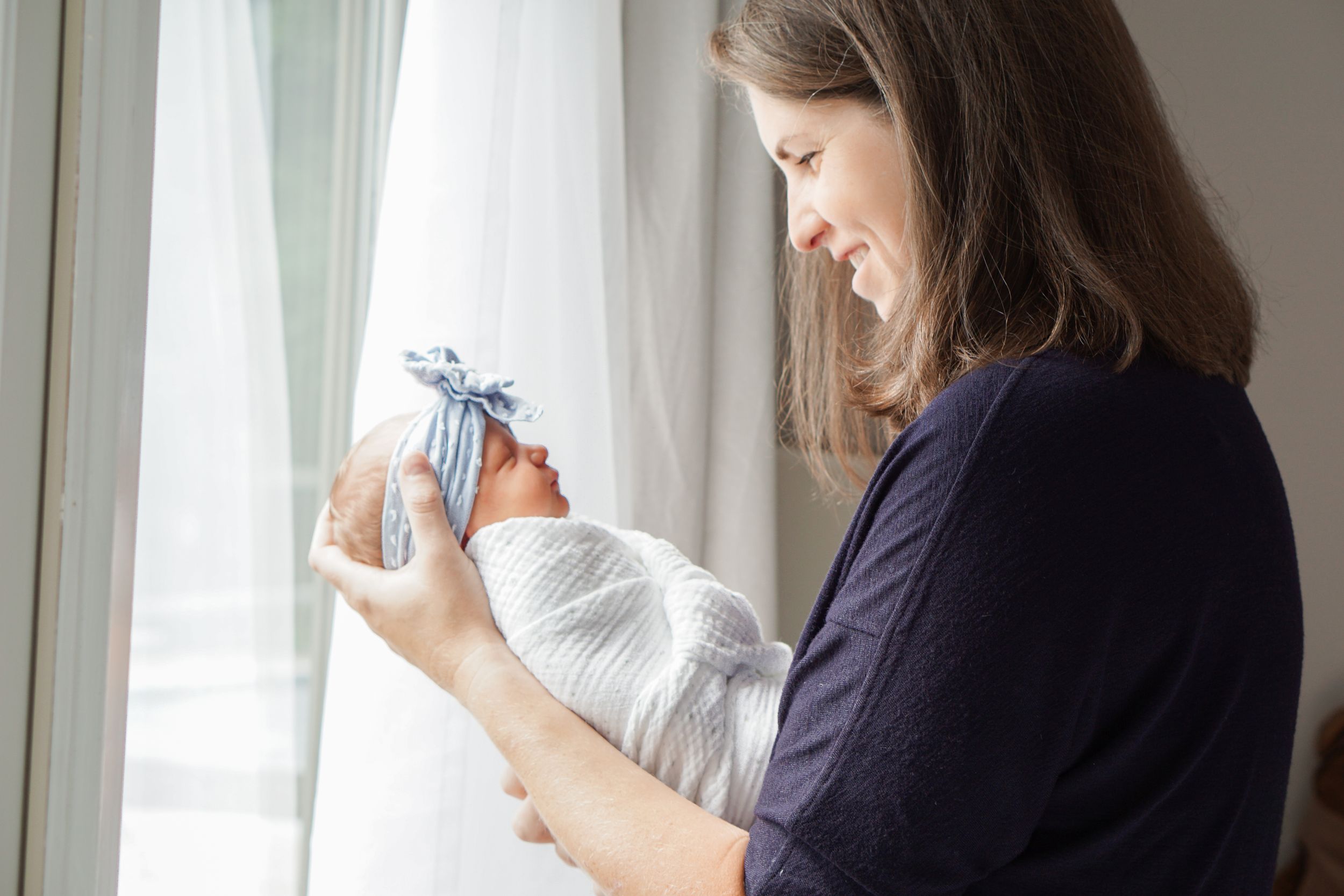A person in navy holds a swaddled infant by a bright window.