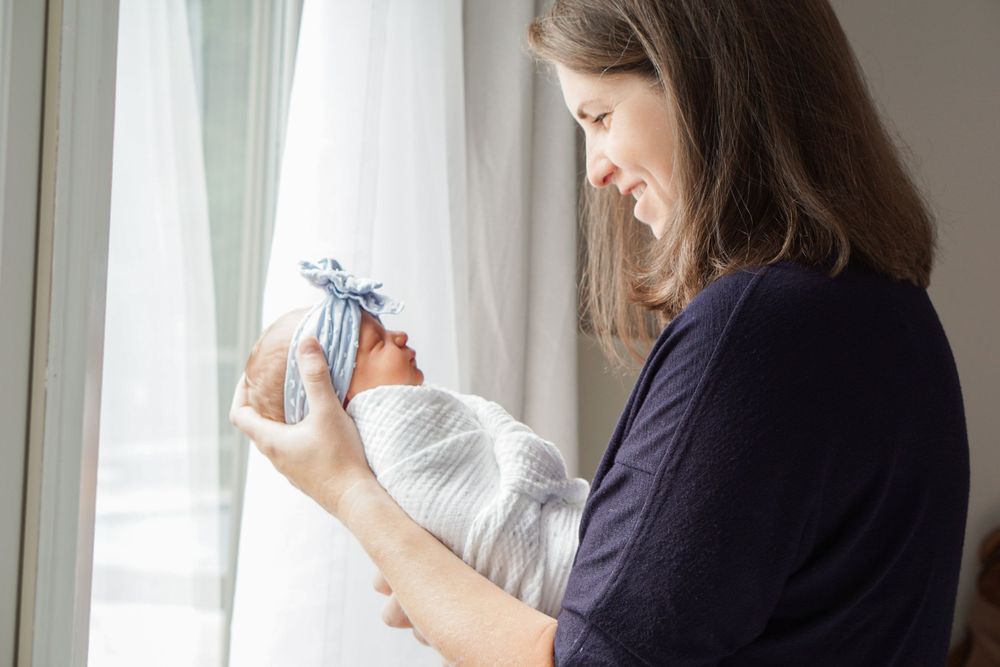 A person in navy holds a swaddled infant by a bright window.