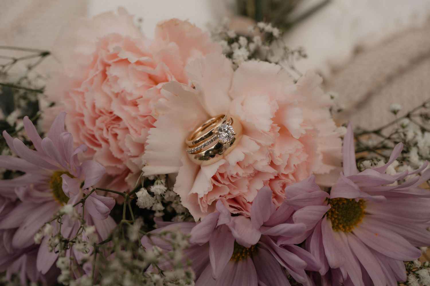 Close-up of pink peonies and purple daisies with wedding rings nestled among the petals.