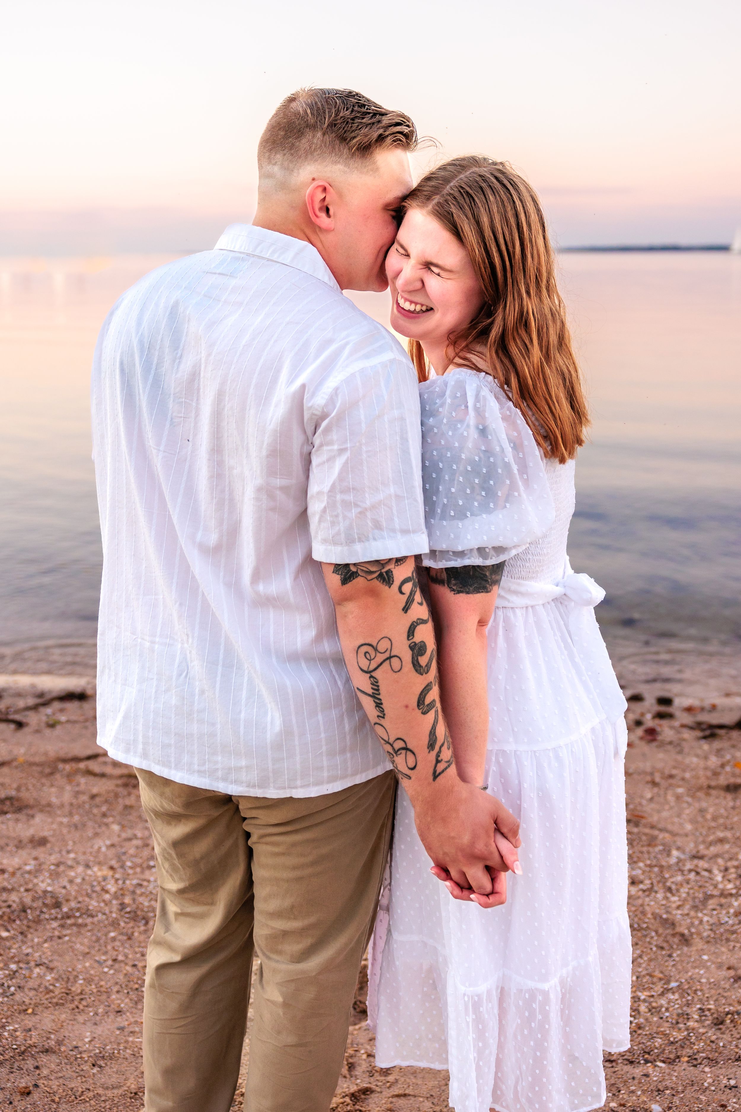 A couple in white outfits sharing an intimate moment on a beach at sunset.