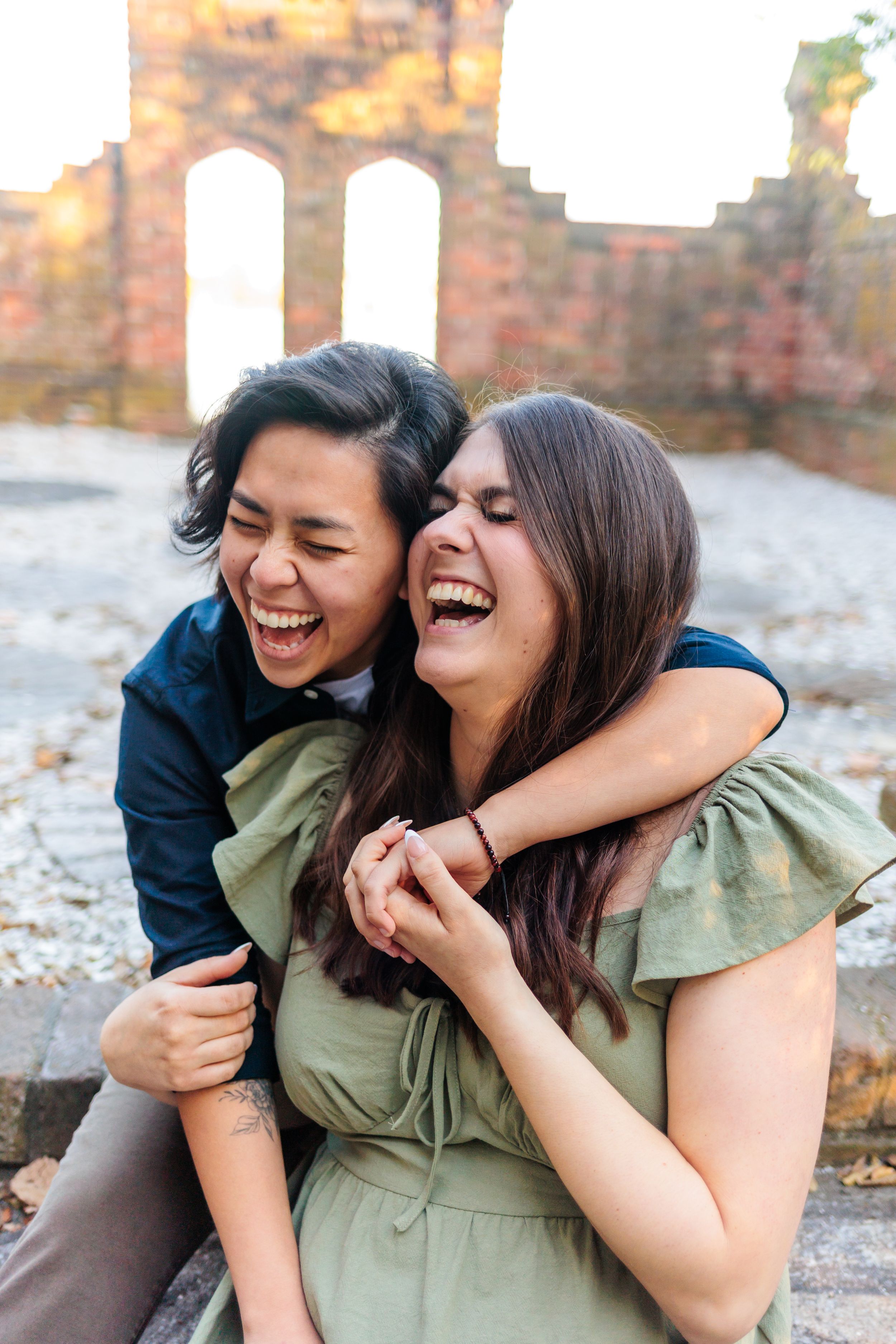 Two people in casual outfits sharing a genuine laugh together against a stone wall backdrop.