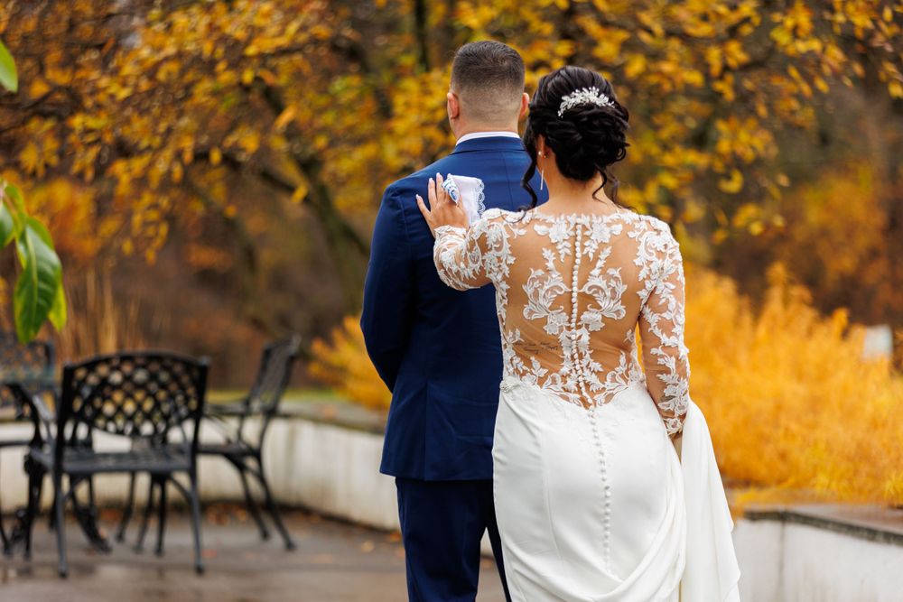 Bride in lace gown and groom in navy suit walking through autumn foliage from behind.